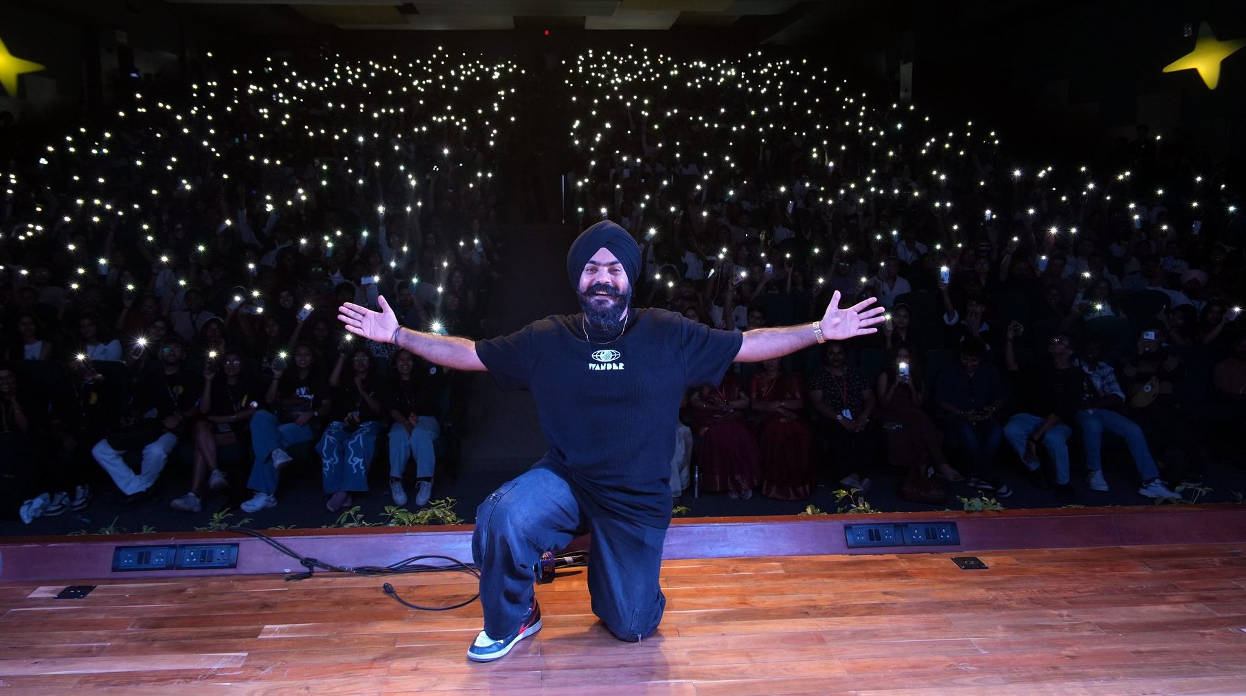 Harman Preet Singh on stage, arms wide, a crowd of phone lights behind him